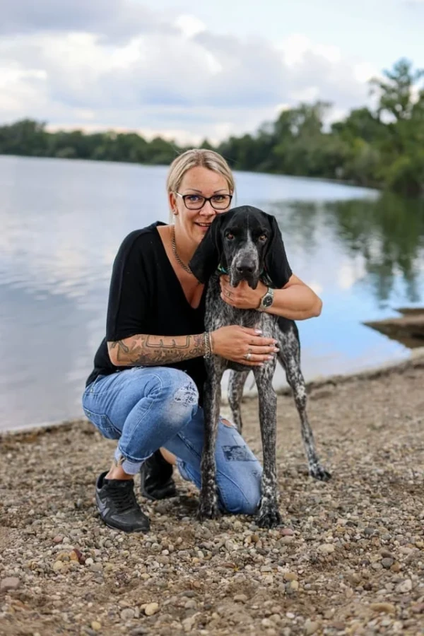 Justine Kabas mit Brille und blondem Haar kniet mit ihrem Hund am See.